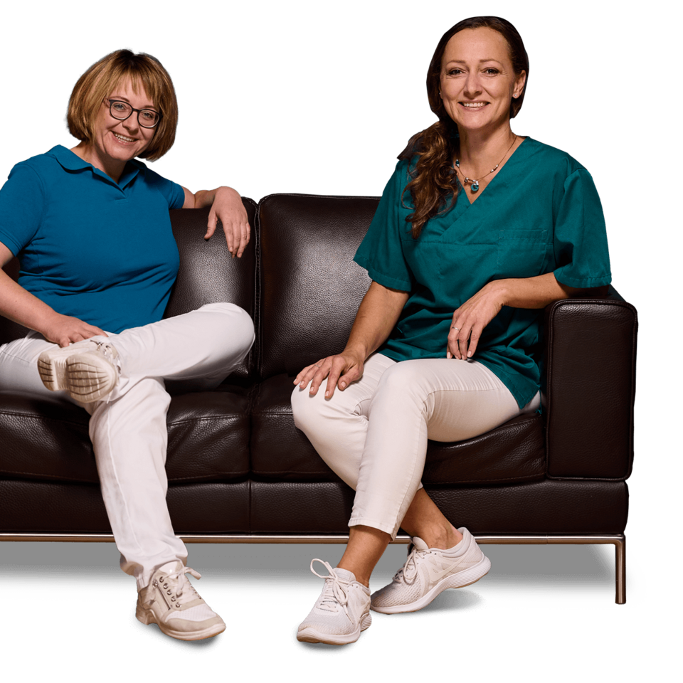 Zahnärztin Dr. Manuela Ringler und ihre Mitarbeiterin Frau Vitztum sitzen nebeneinander auf einer braunen Ledercouch