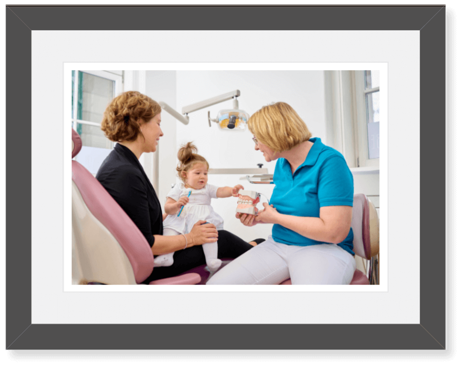 Zahnärztin Dr. Ringler unterhält sich mit einem kleinen Mädchen, das gemeinsam mit seiner Mutter auf dem Behandlungsstuhl sitzt.
