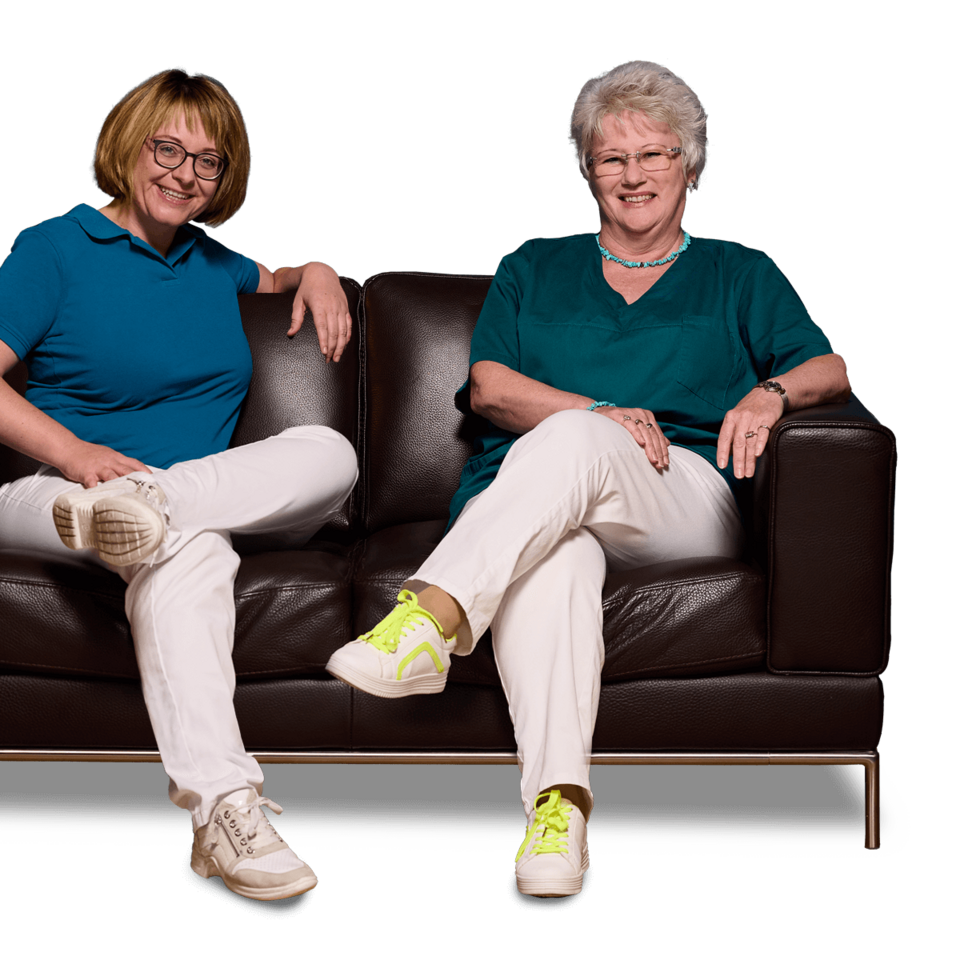 Zahnärztin Dr. Manuela Ringler und ihre Mitarbeiterin Frau Niekisch sitzen nebeneinander auf einer braunen Ledercouch